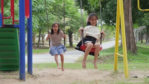 Cute sisters are having fun playing on the swings in a playground in a park Stock Footage 295466061
