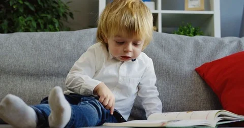 Cute small boy sitting on the couch and looking at the book page through the Stock Footage 82917355