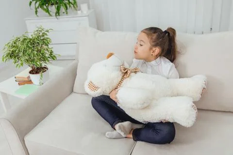 Cute small girl sitting on the sofa in lotos position, lulling her white teddy Stock Photos