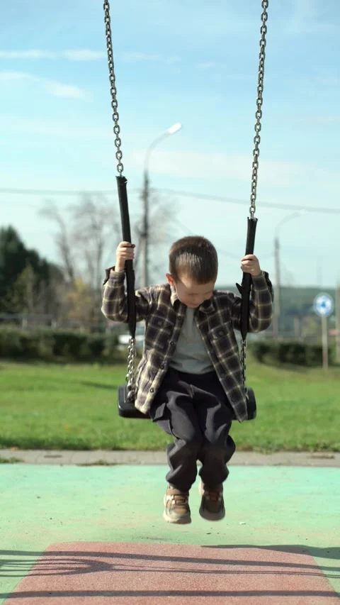 Cute smiling boy sitting on swing at sunny outdoor playground park, holding Vídeo Stock 345528962