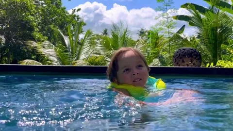 A cute smiling child learns to swim in a beautiful pool surrounded by a tropical 스톡 동영상 272584916