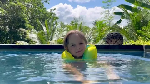 A cute smiling child learns to swim in a beautiful pool surrounded by a tropical 스톡 동영상 272584991