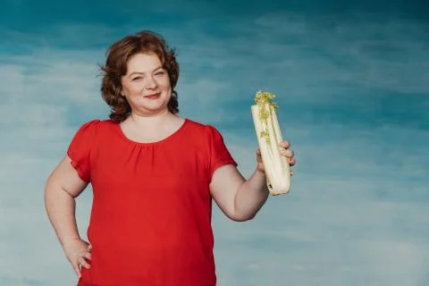 Cute smiling chubby red-haired woman holds out celery against a blue backgrou Stock Photos