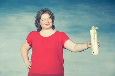 Cute smiling chubby red-haired girl holds out celery on an outstretched arm a Stock Photos