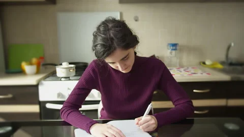 Cute student girl makes notes in a notebook, sitting at the kitchen table Stock Footage 137181302
