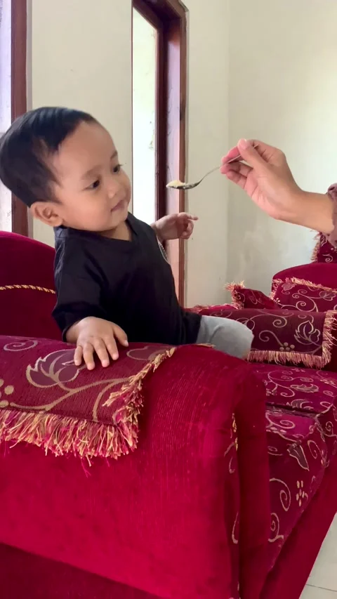 A cute toddler boy playing and climbing on a red velvet sofa in a bright living  库存影片 336843775