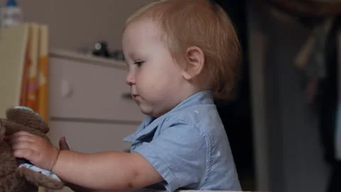 A cute toddler boy playing with a soft brown teddy bear toy in a bright indoor Stock Footage 345610888