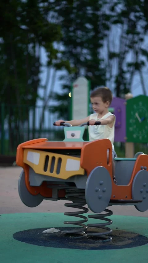 Cute toddler boy sits on bright orange spring-mounted car at outdoor playground, Stock Footage 341410026