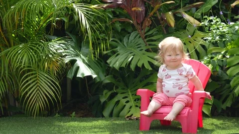 Cute toddler learning to sit on a plastic chair in a lush tropical garden, early Stock Footage 310619903