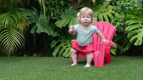 Cute toddler learning to sit on a plastic chair in a lush tropical garden, early Stock Footage 310620152