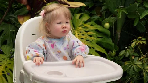 Cute toddler learning to sit on a plastic chair in a lush tropical garden, early Stock Footage 311654150