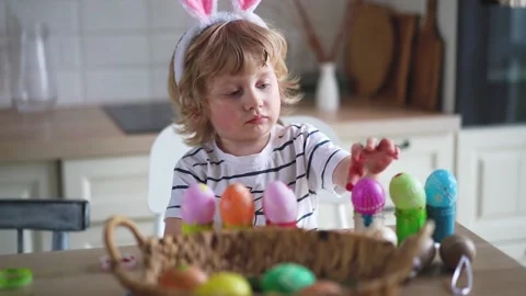 Cute two-year-old boy in bunny ears paint Easter eggs with multi-color paints si Stock Footage 170195342