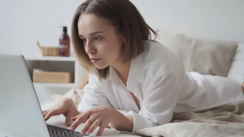 Cute woman is using a laptop while lying in bed and smiling. girl in a man's Stock Footage 125978479