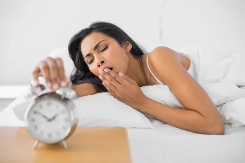 Cute yawning woman lying in her bed while turning off the alarm clock Stock Photos