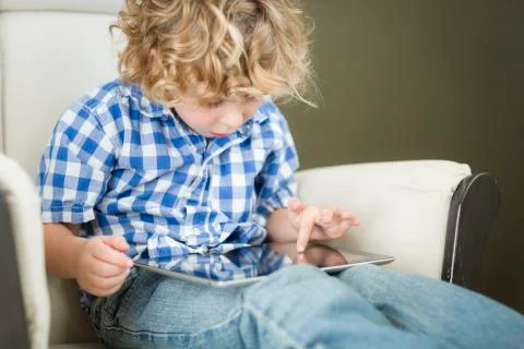 Cute Young Blond Boy Using His Computer Tablet in a Chair. Stock Photos