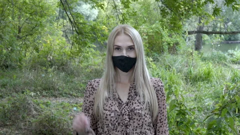A cute young blonde in a black protective mask looks at the camera and waves a 스톡 동영상 140025880