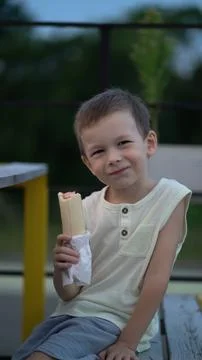 Cute young boy smiling while holding and eating a hot dog outdoors on a woode Stock Photos