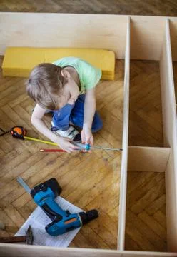 Cute young boy using screwdriver while sitting on floor at unfinished shelf u Stock Photos