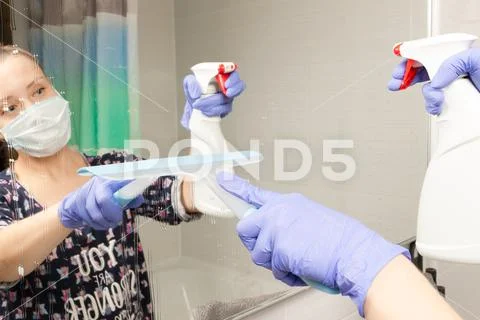 Cute young girl maid in medical mask cleans bathroom mirror during coronaviru Stock Photos