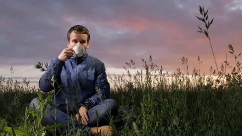 A cute young man drinks a hot drink at sunset on the field. Beautiful sky on a Stock Footage 76991560
