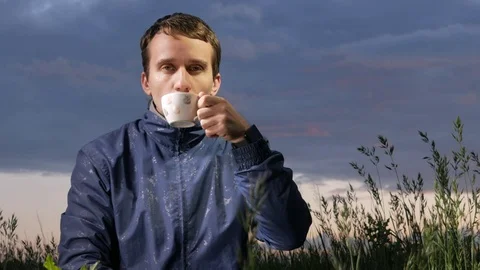 A cute young man drinks a hot drink at sunset on the field. Beautiful sky on a Stock Footage 76991856