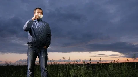 A cute young man drinks a hot drink at sunset on the field. Beautiful sky on a Stock Footage 76992098