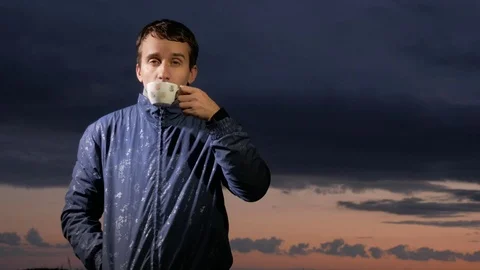 A cute young man drinks a hot drink at sunset on the field. Beautiful sky on a Stock Footage 76992224