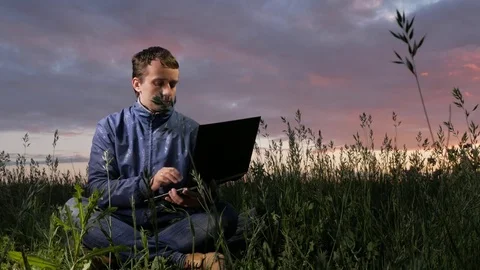 Cute young man working on laptop at sunset on the field. Beautiful sky on a Video stock 76991632