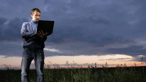 Cute young man working on laptop at sunset on the field. Beautiful sky on a Stock Footage 76992049