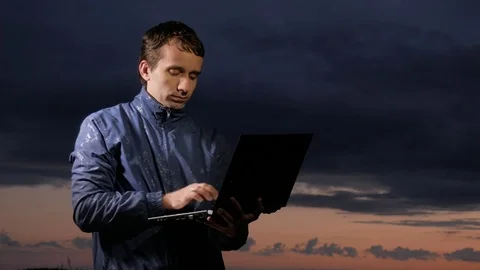 Cute young man working on laptop at sunset on the field. Beautiful sky on a Stock Footage 76992316