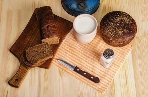 Cutting board, clay pot with milk, rye bread and a knife on a wooden table Stock Photos