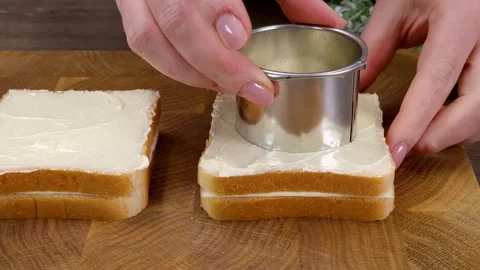 Cutting a round hole in buttered bread slice using a metal cutter on wooden boar Stock Footage 324943237