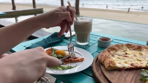 Cutting a slice of pizza with a knife and fork on the beach next to an iced Stock Footage 321561398