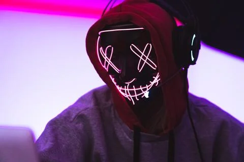 Cyber safety concept. Anonymous hacker in a scary mask wearing headphones and Stock Photos