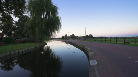 Cyclist approaches along a canal-side path framed by willow trees in rural Stock Footage 346291040