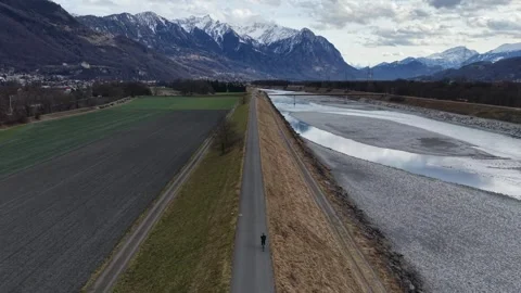 Cyclist Riding on Long Bicycle Path Along River with Mountains Ahead 스톡 동영상 329511651