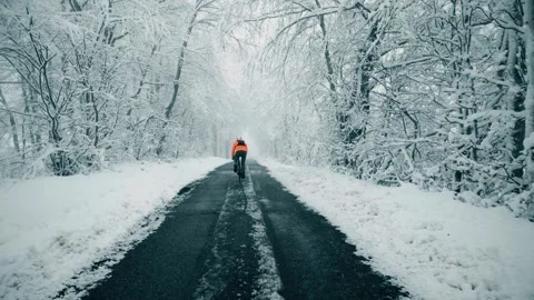 A cyclist wearing orange jacket rides along the forest road in winter as snow 库存影片 315072462