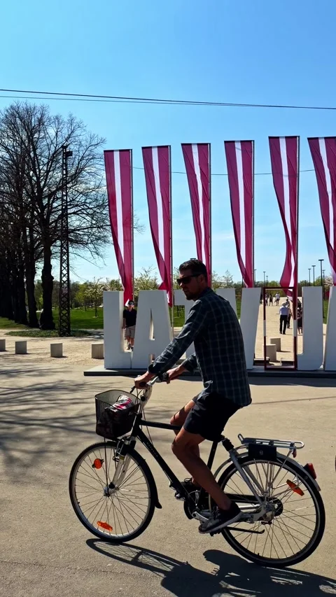 Cyclists and Pedestrians Passing Large LATVIJA Sign in Riga Park on Sunny Stock Footage 335262775