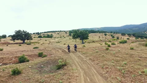 Cyclists ride through a dusty path in a field with hills and trees under a clear Stock Footage 340854715