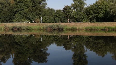 Cyclists riding along a riverside path with lush green trees reflecting in calm  Stock Footage 315159511
