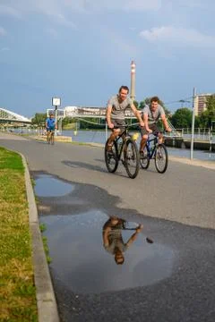 Cyclists on a riverside path with reflection in water puddle Stock Photos