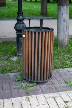 Cylindrical brown slatted trash can with black metal lid.\nSituated on bric.. Stock Photos