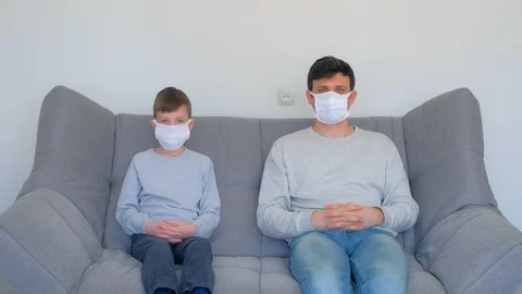 Dad and son in masks sitting on couch at home, coronavirus pandemic quarantine. Stock Footage 128974634