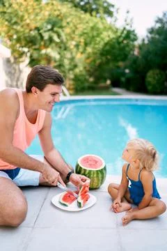 Dad cuts watermelon into slices in front of a little girl sitting by the pool Stock Photos