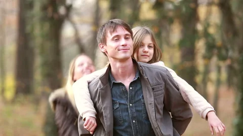 Dad giving daughter piggyback ride, mom throwing leaves at them in park Stock Footage 59554407