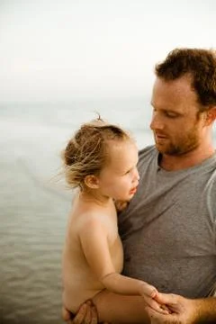 Dad in gray shirt holds naked blonde daughter with sunburn on beach Stock Photos
