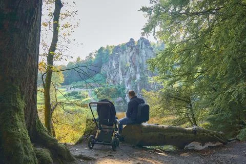 Dad sits on a fallen tree trunk next to a baby in a stroller looking at the lake Stockfoto's