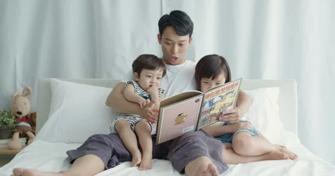 Dad sitting in bed and reading storybook to his son and daughter Stock Footage 100936147