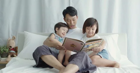 Dad sitting in bed and reading storybook to his son and daughter Stock Footage 100946875