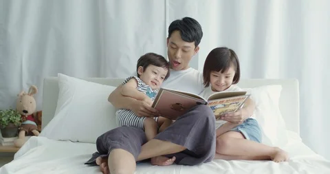 Dad sitting in bed and reading storybook to his son and daughter Stock Footage 100947086